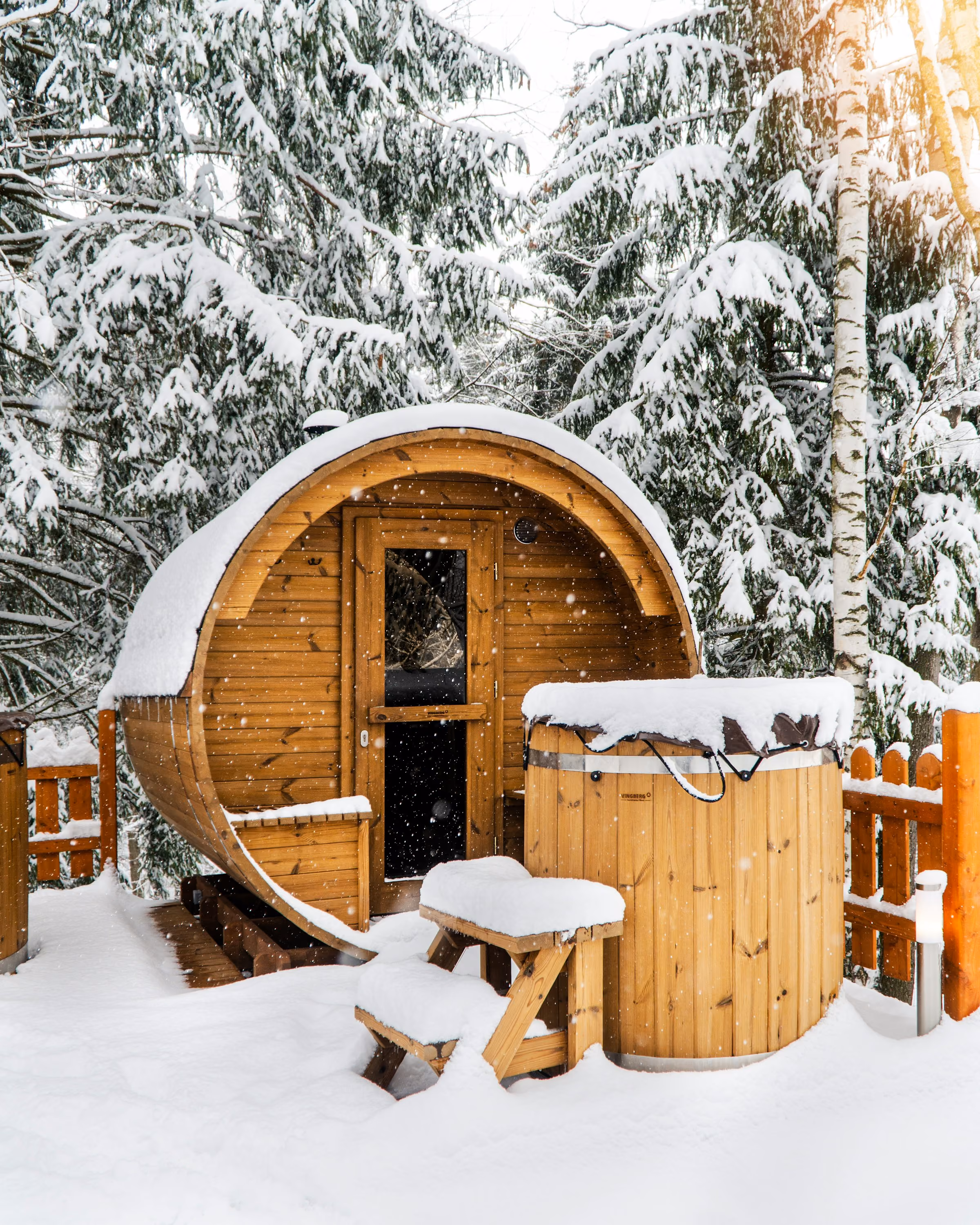 A wooden barrel sauna and hot tub in deep snow among pine trees