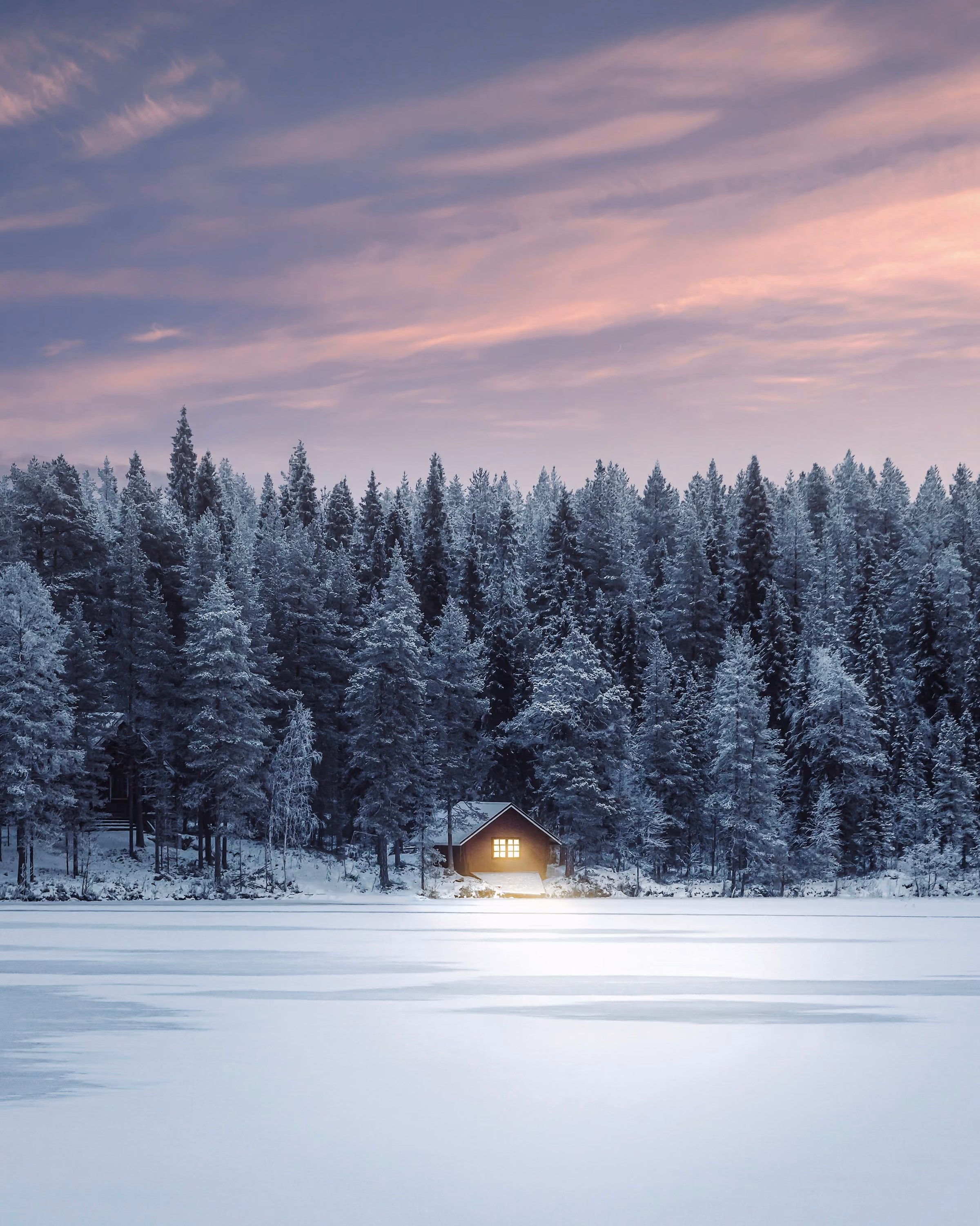 A small lit cabin on the edge of a frozen lake under a pink sky