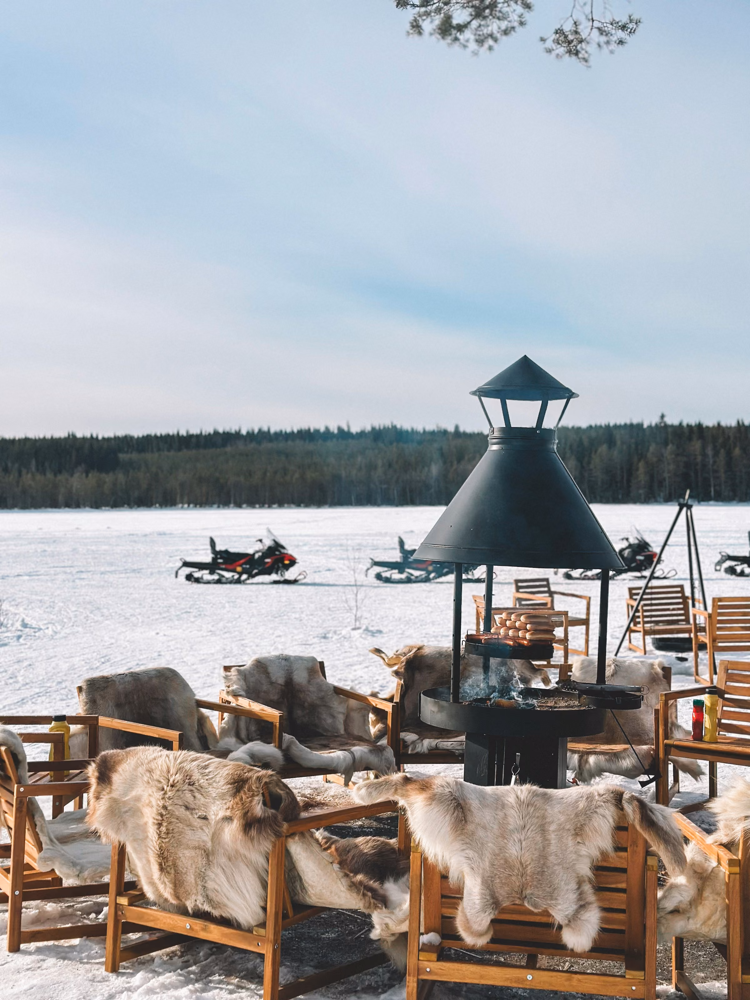 Chairs draped in reindeer hides around a fire under a kota shelter on a frozen lake