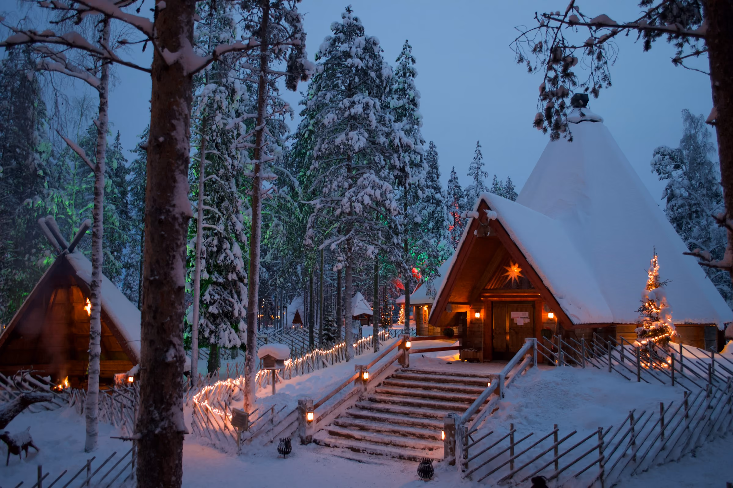 Snow-covered log cabins lit with fairy lights in Santa Claus Village at dusk