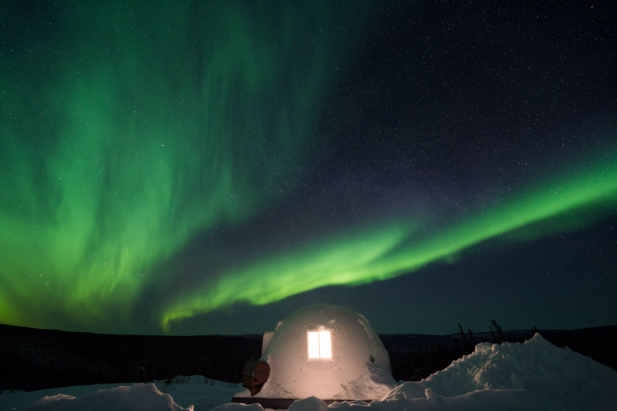 A lit shelter in the snow under a green aurora
