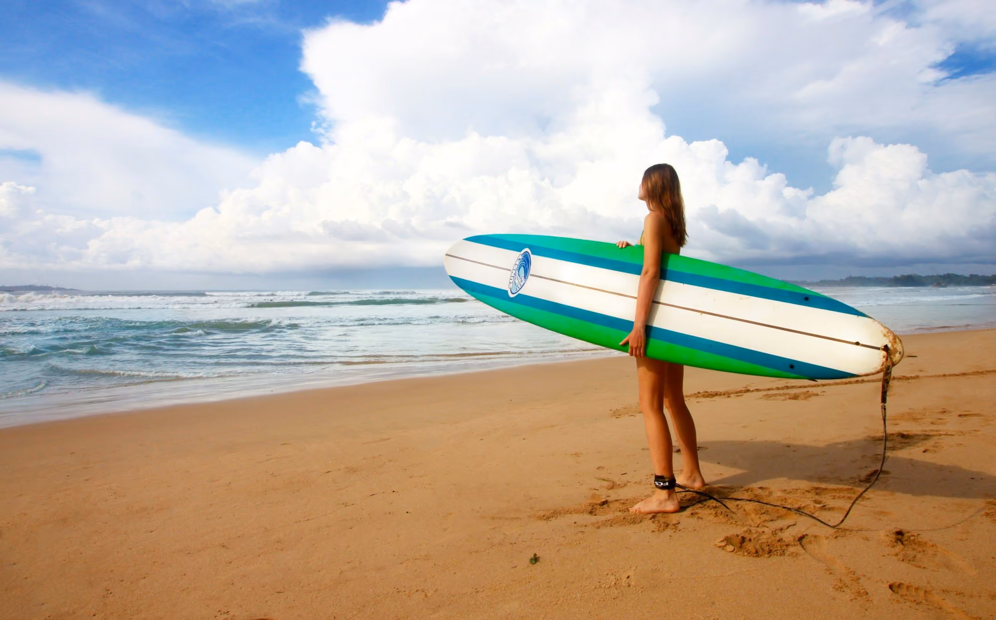 Near Cape Weligama: woman standing near sea holding white, blue, and green surfboard under blue sky