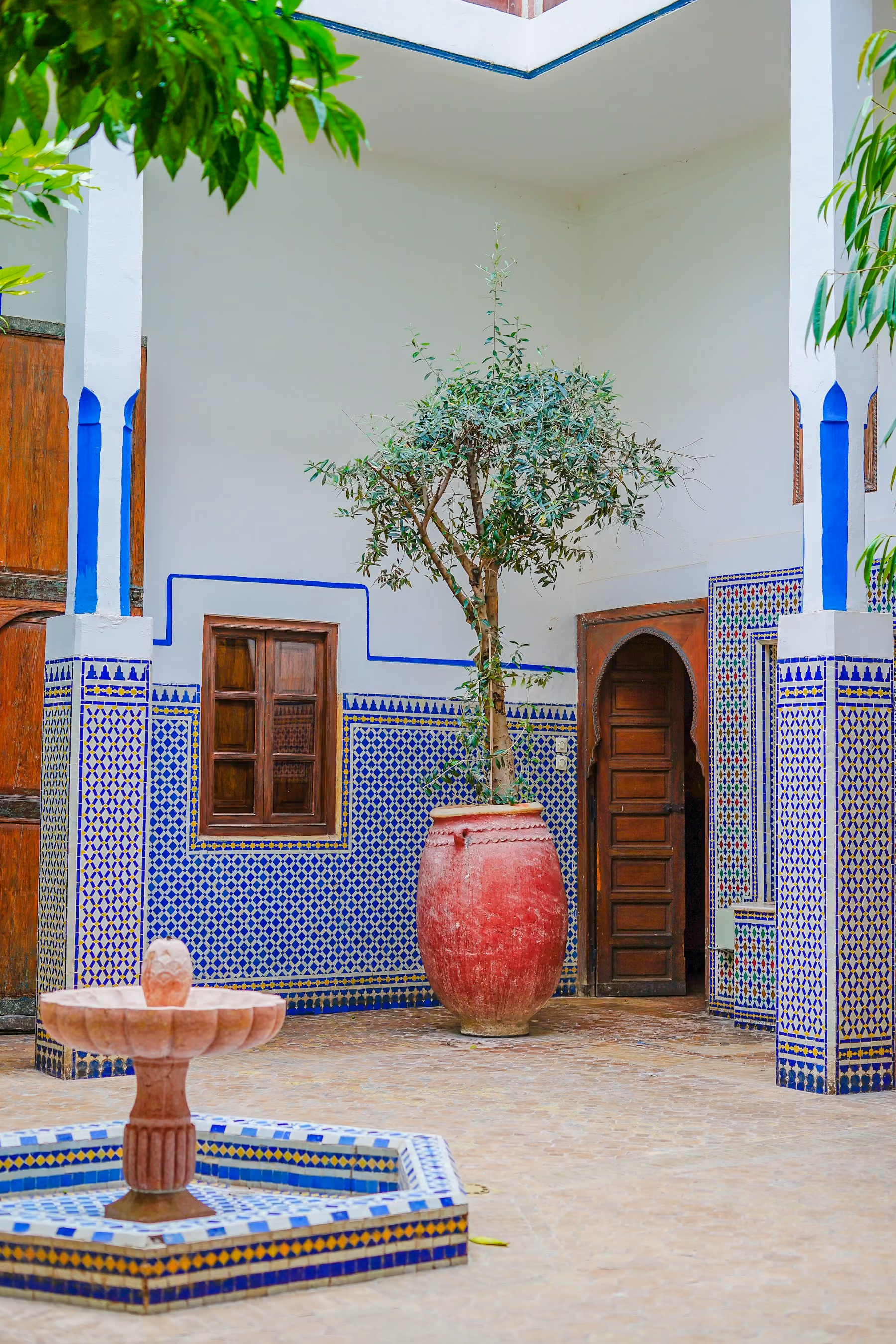 A tiled riad courtyard in Marrakech with a fountain, a potted olive tree and carved wooden doors