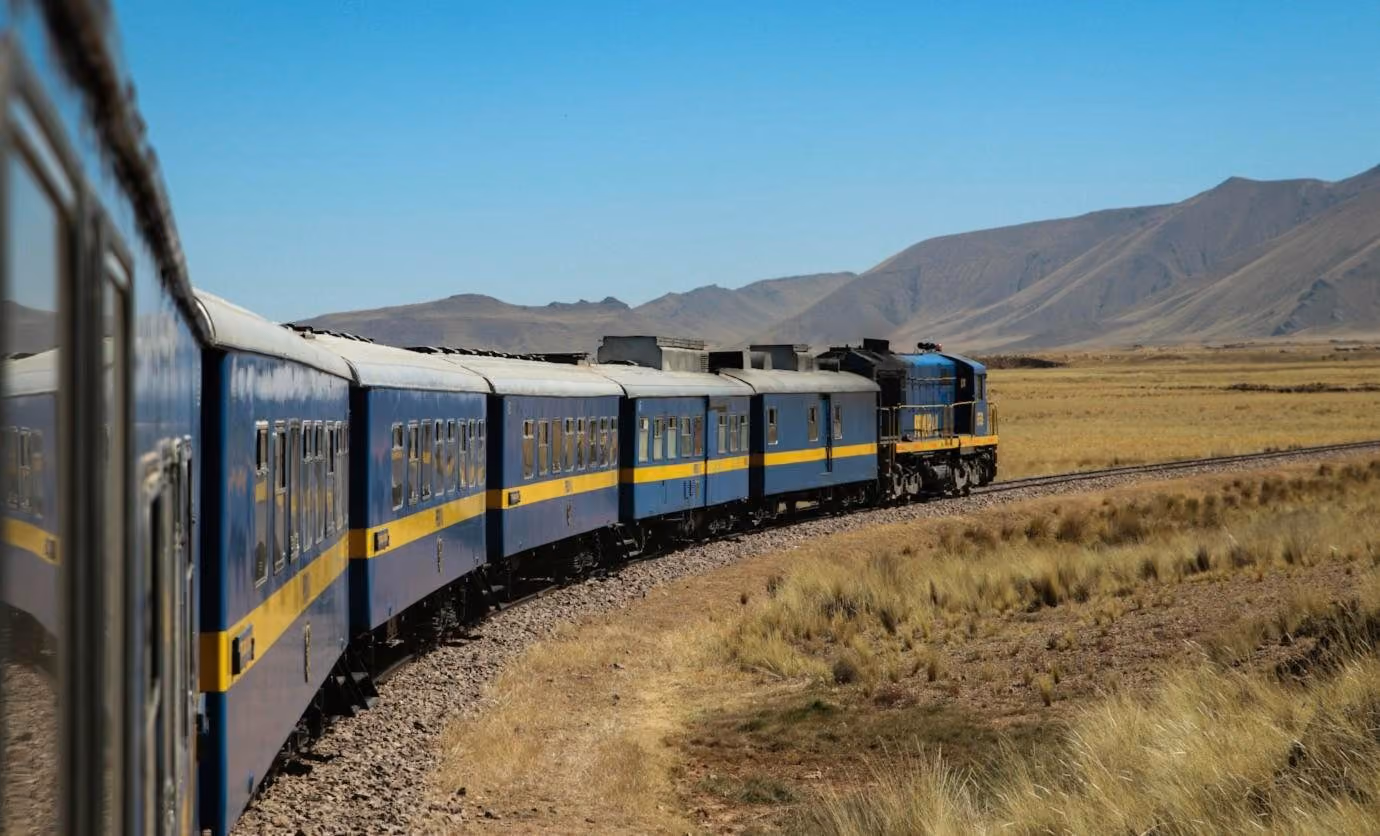 A blue and yellow train curving across the altiplano, mountains behind